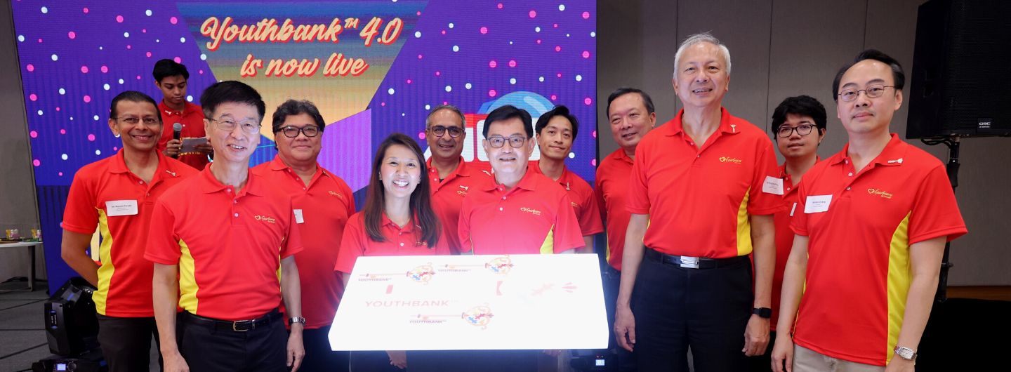 Group of people wearing red Youthbank shirts holding a glowing sign in front of a backdrop.